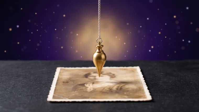 Pendule divinatoire en laiton doré suspendu immobile au-dessus d'une photographie ancienne posée à plat sur une table d'ardoise, fond violet étoilé